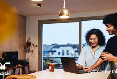 Una mujer y un hombre de pie junto a la encimera de la cocina, mirando una computadora portátil. Están sonriendo.