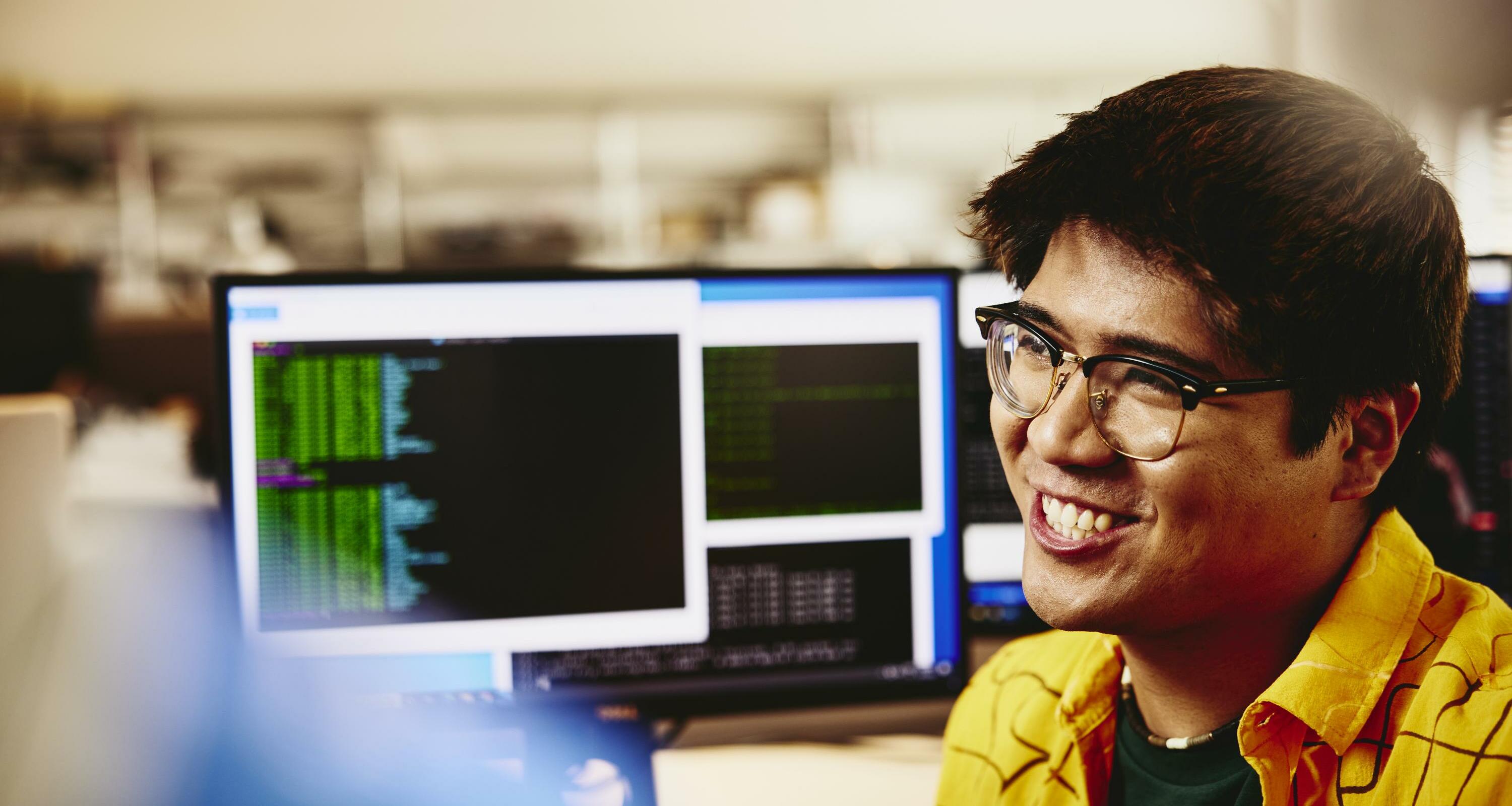 Man smiling while sitting behind his desk, computer screens displaying programming code.
