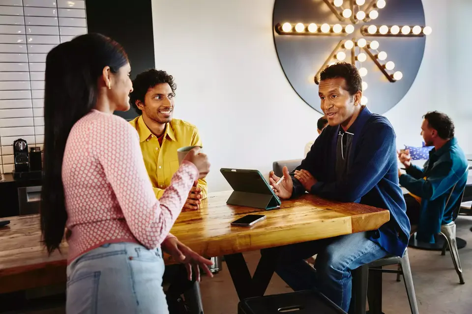 2 male sitting at a table and a female standing at the table with a coffee mug